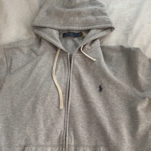 Grå hoodie från Polo Ralph Lauren - Snygg grå hoodie från Polo Ralph Lauren med klassisk blå logga på bröstet. Tröjan har huva med vita snören, dragkedja framtill och två fickor. Mjuk insida i bomullsmix som gör den extra skön. Perfekt för chill dagar eller när du vill ha en clean look.