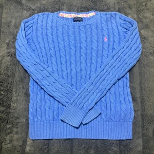 Blå kabelstickad tröja Polo Ralph Lauren - Kabelstickad blå tröja från Polo Ralph Lauren i storlek XL(16 år). Tröjan har långärmad design, ribbade muddar och rund hals. På bröstet finns en rosa broderad logga. Vid frågor är det bara skriva