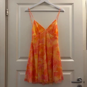 Färgglad kort klänning med snörning - Superfin kort klänning från Zara i orange, rosa och gul med blommigt mönster. Klänningen har tunna axelband, v-ringning och snygg snörning i ryggen. Perfekt för sommarens varma dagar och har en flowy passform som känns riktigt luftig. aldrig använd! Prislapp är kvar