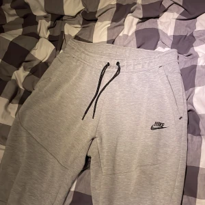 Grå mjukisbyxor från Nike, storlek M - Snygga grå mjukisbyxor från Nike med svart logga på benet. Byxorna har svarta snören i midjan och dragkedja på bakfickan. Tillverkade i mjukt material som är perfekt för chill eller träning. Klassisk passform och stilren look.