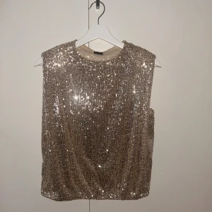 Guldig paljettopp med axelvaddar - Säljer en glittrig guldig topp täckt av paljetter. Modellen är ärmlös med breda axlar och axelvaddar för en cool siluett. Perfekt för att sticka ut och fånga ljuset. Rund halsringning och rak passform. Superfin till svarta byxor eller kjol.