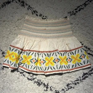 Broderad kortkjol med blommor - Söt kortkjol i ljusbeige bomull med broderade blommor i gult och orange samt gröna blad. Kjolen har smock upptill med zigzag-mönster i grönt och orange, och volang nedtill. Perfekt för dig som gillar boho-vibe och färgglada detaljer.