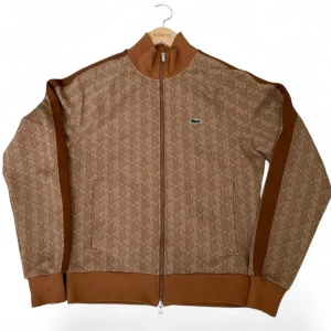 Brun mönstrad cardigan från Lacoste - Säljer en brun cardigan från Lacoste med diskret geometriskt mönster och klassisk krokodillogga på bröstet. Jackan har ribbade muddar, hel dragkedja och bruna paneler längs ärmarna. Perfekt för dig som gillar sportig och stilren design. Pris kan diskuteras!