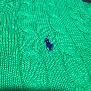 Grön stickad tröja från Polo Ralph Lauren - Snygg grön kabelstickad tröja från Polo Ralph Lauren i storlek S. Tröjan har rund hals, långärmade ribbade muddar och klassisk blå broderad logga på bröstet. Perfekt för dig som gillar preppy stil och vill ha något som sticker ut.