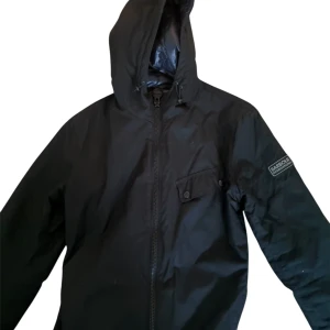 Svart fodrad jacka från Barbour M - Svart jacka från Barbour International med huva och dragkedja framtill. Jackan har en ficka med knapp på bröstet och Barbour-logga på ärmen. Materialet är syntet och jackan är fodrad för extra komfort. Perfekt för blåsiga dagar. Nypris  3500