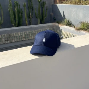 Mörkblå keps från Polo Ralph Lauren - Snygg mörkblå keps från Polo Ralph Lauren med klassisk vit broderad logga framtill. Kepsen har böjd skärm, justerbar rem baktill i metall och är tillverkad i mjuk bomull. Perfekt för att lyfta din streetstyle eller sportiga look. vet inte om den är äkta
