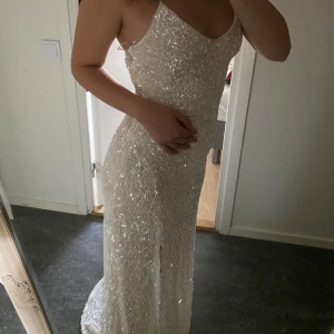 Glittrig långklänning med slits - Supersnygg långklänning i glittrigt material med tunna axelband och djup urringning. Klänningen är täckt av paljetter och har en hög slits framtill som ger en extra wow-effekt. Perfekt för dig som vill sticka ut och glänsa på bal eller fest. Passar som bröllopsklänning därmed. Klänningen är helt ny och endast testad på bilden, prislappen finns kvar 