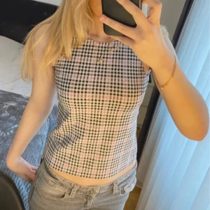 Rutig ärmlös linnetopp i rosa och svart - Snygg ärmlös linnetopp med rutigt mönster i svart, vitt och ljusrosa. Toppen har rund halsringning och dragkedja i ryggen. Perfekt till jeans eller kjol för en trendig look. Den är i barn storlek, men skulle säga att det sitter som en XS på mig. Har ej använts.