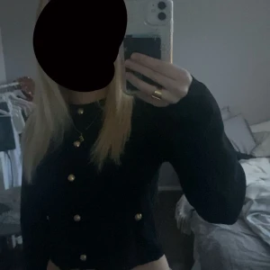 Svart kofta med guldknappar - Snygg svart kofta med guldiga knappar framtill. Modellen är croppad och har långa ärmar. Perfekt för dig som vill ha en stilren men ändå trendig look.