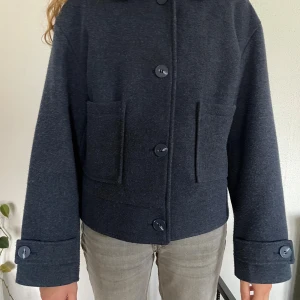 Mörkblå ulljacka med stora knappar - Säljer en mörkblå, rak och lite boxig ulljacka med stora knappar framtill och på ärmsluten. Jackan har två rejäla fickor fram och är långärmad. Perfekt för dig som gillar en clean och stilren look.