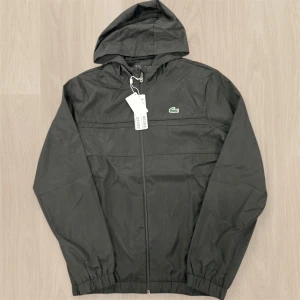 Svart Lacoste Sport kofta - Svart vindjacka från Lacoste Sport med huva och dragkedja framtill. Jackan har elastiska muddar, meshfoder och den klassiska gröna krokodilloggan på bröstet. Perfekt för dig som vill ha en clean och sportig look.