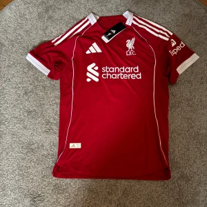 Liverpool röd fotbollströja Adidas Wirtz 7 - Liverpool FC matchtröja i rött med vita detaljer från Adidas. Kortärmad modell med klubbmärke, sponsortryck och spelarnamn Wirtz samt nummer 7 på ryggen. Klassiska tre ränder på axlarna och vit krage. Tillverkad i lätt och ventilerande material.