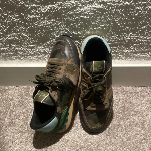 Valentino rockrunner - Säljer ett par Valentino rockrunner sneakers med camouflage-mönster i grönt, brunt och svart. Skorna har detaljer i mocka, skinn och textil samt en cool turkos detalj vid hälen. Klassisk snörning och robust sula. Perfekta för dig som vill sticka ut med lyxiga och unika sneakers. Själv klart äkta ✅