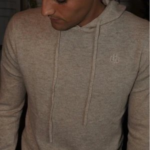 Beige  kashmere hoodie från Bvaldi med luva  - Mjuk och skön beige hoodie från Bvaldi med huva och snörning. Tröjan är stickad och har långa ärmar samt ribbade muddar vid ärmslut och nederkant. Perfekt för dig som gillar enkel och clean stil. Tröjan är oanvänd 