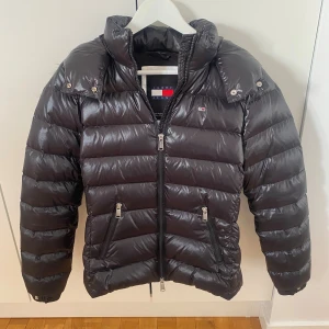 Svart pufferjacka från Tommy Jeans - Svart, glansig pufferjacka från Tommy Jeans med avtagbar huva och snygg fuskpälsdetalj baktill. Jackan har två fickor med dragkedja, hög krage och quiltad design. Perfekt för kalla dagar och har en modern, loose passform.