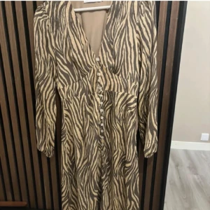 Zebramönstrad långklänning med knappar - Snygg långklänning med zebramönster i beige och svart. Klänningen har v-ringning, långa ärmar och knappar framtill. Materialet är lätt och har en krinklad struktur som ger en cool känsla. Perfekt för dig som vill sticka ut med ett djurmönster. Två knappar är borta men inget som syns och går att köpa.❤️Pris kan diskuteras vid snabb affär.❤️