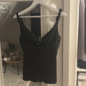 Svart linnetopp från Adoore - Säljer en svart linnetopp från Adoore med snygg draperad urringning och twistade axelband. Toppen har en figurnära passform och är tillverkad i ett slätt, lätt glansigt material som ger en lyxig känsla. Perfekt att styla med jeans eller kjol.