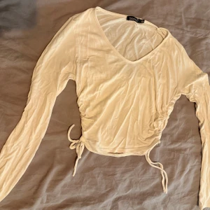 Vit/Beige croppad topp från Boohoo - Säljer en vit/beige långärmad croppad topp från Boohoo med v-ringning och snygg snörning i sidorna. Tunt och mjukt material.