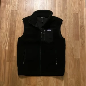 Svart fleeceväst från Patagonia L - Svart fleeceväst från Patagonia i storlek L sitter som en medium. Västen har hög krage, hel dragkedja framtill och tre fickor med dragkedja. Klassisk och stilren design med diskret logga på bröstet. Perfekt lager-på-lager-plagg för kyliga dagar.