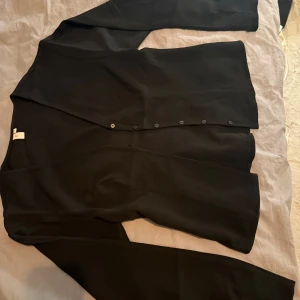 Svart kofta från Nelly med knappar - Oanvänd. Svart stilren kofta från Nelly i storlek S. Koftan har långa ärmar och knappar framtill, perfekt för lager-på-lager. Materialet är som en kavaj. Enkel och clean design som funkar till många olika looks. 