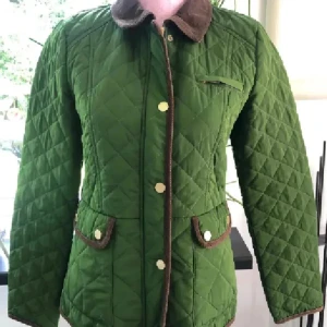 Grön quiltad jacka från Stockh LM - Quiltad grön jacka från Stockh LM med bruna manchesterdetaljer på krage och fickor. Jackan har guldiga knappar, figursydd passform och snygga detaljer baktill. Perfekt för dig som gillar klassisk stil med modern twist.