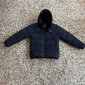 Mörkblå pufferjacka från BOSS - Snygg mörkblå pufferjacka från BOSS med huva och diskret logga på bröstet och ärmen. Jackan har quiltad design, dragkedja framtill och två sidofickor. Perfekt för kalla dagar och har en clean, modern look som funkar till streetwear.
