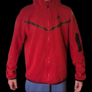 Röd Nike fleecejacka med dragkedja - Nike fleecejacka i rött med svart detalj över bröstet och logga på vänster sida. Jackan har huva, hel dragkedja och ficka med dragkedja på ärmen. Svarta muddar vid ärmslut och nederkant. Perfekt för chill eller när du vill ha något mjukt och skönt.