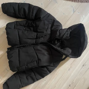Svart pufferjacka med huva - Svart pufferjacka från Zalando. Köpt för nått år sedan och har inte använt på ett tag. Dragkedjan är lite defekt och är ganska svår att stänga/öppna. Det går om man verkligen vill annars är jackan snygg som öppen. 