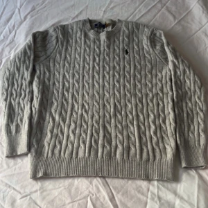 Grå kabelstickad tröja Polo Ralph Lauren - Kabelstickad grå tröja från Polo Ralph Lauren med klassisk rund halsringning och broderad svart logga på bröstet. Tröjan är i mjuk ullblandning och har ribbade muddar vid ärmslut och nederkant. Perfekt för lager-på-lager under kyliga dagar. klädlängd 68 och axellängd 42