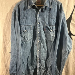Levi's jeansskjorta XXL med pärlknappar - Vintage Klassisk ljusblå jeansskjorta från Levi's i storlek XXL. Skjortan har två bröstfickor med lock och pärlknappar, samt tydliga western-detaljer. Tillverkad i bomull och har långa ärmar. Hål i båda armhålorna som inte syns när man har på sig den, den passar som en XL