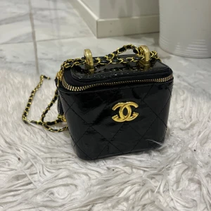 Svart väska - Supersnygg svart glansigt skinn med ikoniskt CC-emblem i guld. Väskan har kedjerem med gulddetaljer och dragkedja upptill. Perfekt för dig som vill ha en lyxig och trendig accessoar med exklusiv känsla.