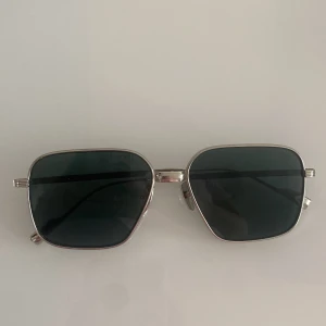 Silvriga CHIMI solglasögon fyrkantiga - Snygga solglasögon från CHIMI med fyrkantiga, mörka linser och silvriga metallbågar. Modern design med stilrena detaljer och bekväma näskuddar. Perfekt accessoar för dig som vill ha en cool och tidlös look.