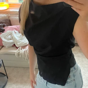 Svart asymmetrisk blus - Säljer en svart blus med snygg asymmetrisk halsringning och ärmlös design. Toppen har en stilren och minimalistisk look som passar till jeans eller kjol. Materialet känns mjukt och har en lätt glans, vilket ger en exklusiv känsla.