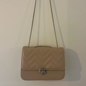 Beigerosa quiltad axelväska med kedja - Snygg beige axelväska med quiltat mönster och silverfärgad kedjerem. Väskan har ett stilrent spänne framtill och är tillverkad i skinnimitation. Perfekt storlek för det viktigaste och ger en trendig touch till din outfit.