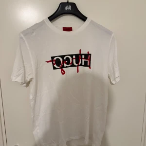 Vit HUGO t-shirt med logga - Vit t-shirt från HUGO med svart och röd logga tryckt på bröstet. Klassisk rund halsringning och korta ärmar. Tillverkad i mjuk bomull för en skön känsla och clean look. Äkta såklart