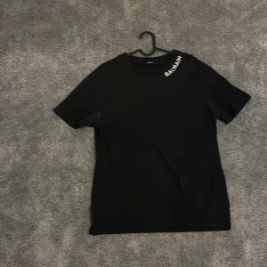 Svart t-shirt från Balmain Paris - Svart t-shirt från Balmain Paris med vit logga tryckt vid halsen. Klassisk rund halsringning och korta ärmar. Tillverkad i mjuk bomull för en skön känsla och clean look. Perfekt för dig som gillar stilrena designer och exklusiva märken.