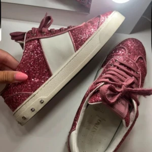 Valentino Garavani rosa glittriga sneakers - Säljer ett par Valentino Garavani sneakers i rosa med glittrigt mönster och vita detaljer. Skorna har snörning, platt sula och små nitar på sidan. Insidan är vit med logotyp. Perfekt för dig som vill sticka ut med coola och lyxiga sneakers. i storlek 39 extra snören i vit och samt kartorn till skorna 