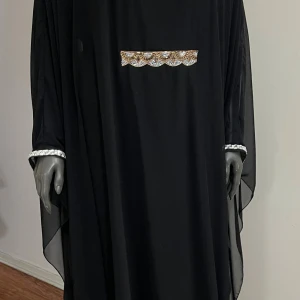 Svart långklänning med gulddetaljer - Elegant svart långklänning med löst fall och vackra guldfärgade broderier vid halsringningen och på bröstet. Ärmarna är vida och dekorerade med glittriga detaljer vid mudden. Klänningen har en flowy siluett och känns riktigt lyxig.