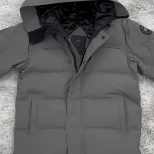 Canada goose MACMILLAN - Snygg grå pufferjacka från Canada Goose med huva och svart patch på ärmen. Jackan har två sidofickor, dragkedja med logga och knappar framtill. Insidan är fodrad i svart och materialet är slitstarkt och vattenavvisande, perfekt för kalla dagar. Storlek S. Jackan användes i Februari förra året fick den som present i min födelsedag. Har växt mycket sedan dess. Vill verkligen sälja den snabbt därav det låga priset du kan sälja denna för dubbla priset Minst.