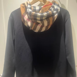 Svart Burberry hoodie med rutig huva - Svart hoodie från Burberry i storlek M med klassisk rutig huva i beige, brun och svart. Tröjan har dragkedja framtill, snörning vid huvan och två fickor. Perfekt för dig som gillar stilrena streetwear-plagg med lyxig touch.
