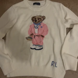 Vit stickad tröja Polo Ralph Lauren - Vit stickad tröja från Polo Ralph Lauren med ikonisk björn i rosa outfit broderad på bröstet. Rund halsringning, ribbade muddar och RL-broderi nertill. Perfekt statement-piece för dig som gillar klassisk stil med en twist.
