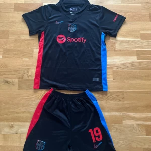 FC Barcelona Lamine Yamal fotbollströja - Svart FC Barcelona fotbollströja med Lamine Yamal och nummer 19 på ryggen. Tröjan har korta ärmar, röda och blå detaljer på sidorna, samt klubbmärke och sponsortryck. Materialet är Dri-FIT ADV polyester som andas. Matchande shorts med samma färgtema ingår. Tröjan är storlek 28. Shorts ingår. Tröja/shorts är i nyskick. Använd en enstaka gång. Passar cirka 10-12 år. 