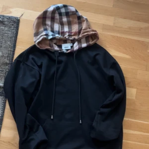 Burberry svart hoodie med rutig huva - Svart hoodie från Burberry i storlek M med klassisk rutig huva i beige, svart och rött. Tröjan har dragsko vid huvan och en stor magficka. Perfekt för dig som vill ha en stilren men ändå unik look. Passar S-M