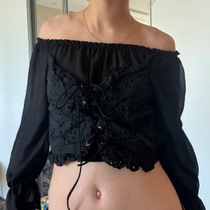 Svart offshoulder korsettopp med snörning - Säljer en svart offshoulder korsettopp med långa, transparenta ärmar och snygg snörning framtill. Toppen är croppad och har broderade detaljer och hålmönster på livet. Perfekt för dig som vill sticka ut med en edgy och romantisk vibe. Står varken storlek eller märke men gissar runt xs/s och köpt i England. Inte från zara!
