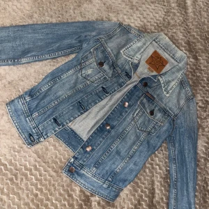 Blå jeansjacka från Gul&Blå XS - Säljer en klassisk blå jeansjacka från Gul&Blå i storlek XS. Jackan har snygga metallknappar framtill, två bröstfickor med lock och en cool läderpatch med logga på insidan. Perfekt för dig som gillar retrovibbar och tidlös stil.