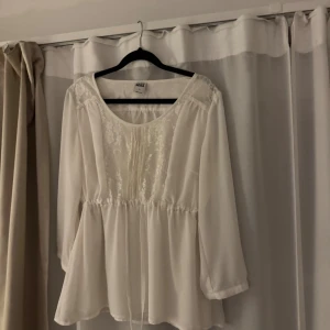 Vit spetsblus från Vero Moda - Supersöt vit blus från Vero Moda med spetsdetaljer framtill och längs axlarna. Blusen har rund halsringning, långa ärmar och dragsko i midjan för en snygg passform. Perfekt för dig som gillar romantisk och feminin stil.