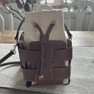 Brun axelväska i skinn och linne - Snygg axelväska med brunt skinn och detaljer i beige linne. Väskan har en unik design med breda skinnremmar och snörning upptill. Justerbar axelrem och guldiga metalldetaljer. Perfekt för dig som vill ha en trendig och stilren accessoar.