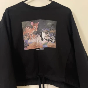 Svart Disney Bambi tröja Pull&Bear - Svart långärmad tröja från Pull&Bear x Disney med tryck av Bambi och Stinka på bröstet samt texten 'Perfect Strangers'. Tröjan är croppad med snörning nertill och har en avslappnad passform. Perfekt för dig som älskar Disney och vill ha en unik streetstyle.