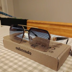 Oversized solglasögon från Cabecade - helt nya i box - Snygga solglasögon från Cabecade med stora, kantiga svarta bågar och guldiga detaljer. Glasen är tonade i mörkgrått och ger en cool vibe. Metallbåge upptill och svarta skalmar med gulddetaljer för en extra lyxig känsla.