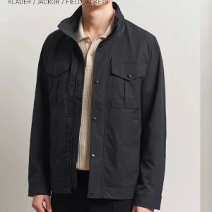 Svart vindjacka med fickor - Stilren svart vindjacka med hög krage och flera praktiska fickor framtill. Jackan har knappar och dragkedja, perfekt för dig som vill ha en clean look med funktionella detaljer. Passar till många olika outfits och har en modern, rak passform.